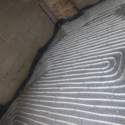 HYDROMAR - Szare rury ogrzewania podłogowego ułożone spiralnie na czarnej macie izolacyjnej, widoczne na tle surowego betonowego stropu i fragmentu ceglanego muru.