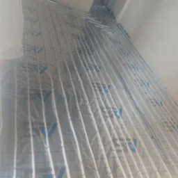 Białe rurki ogrzewania podłogowego ułożone na srebrnej folii z niebieskim nadrukiem, widok z góry, instalacja w trakcie realizacji.