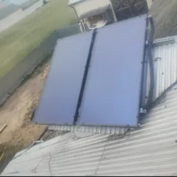 Dwa panele solarne zamontowane na blaszanym dachu budynku, widoczne przewody instalacji. W tle trawnik i budynki gospodarcze.