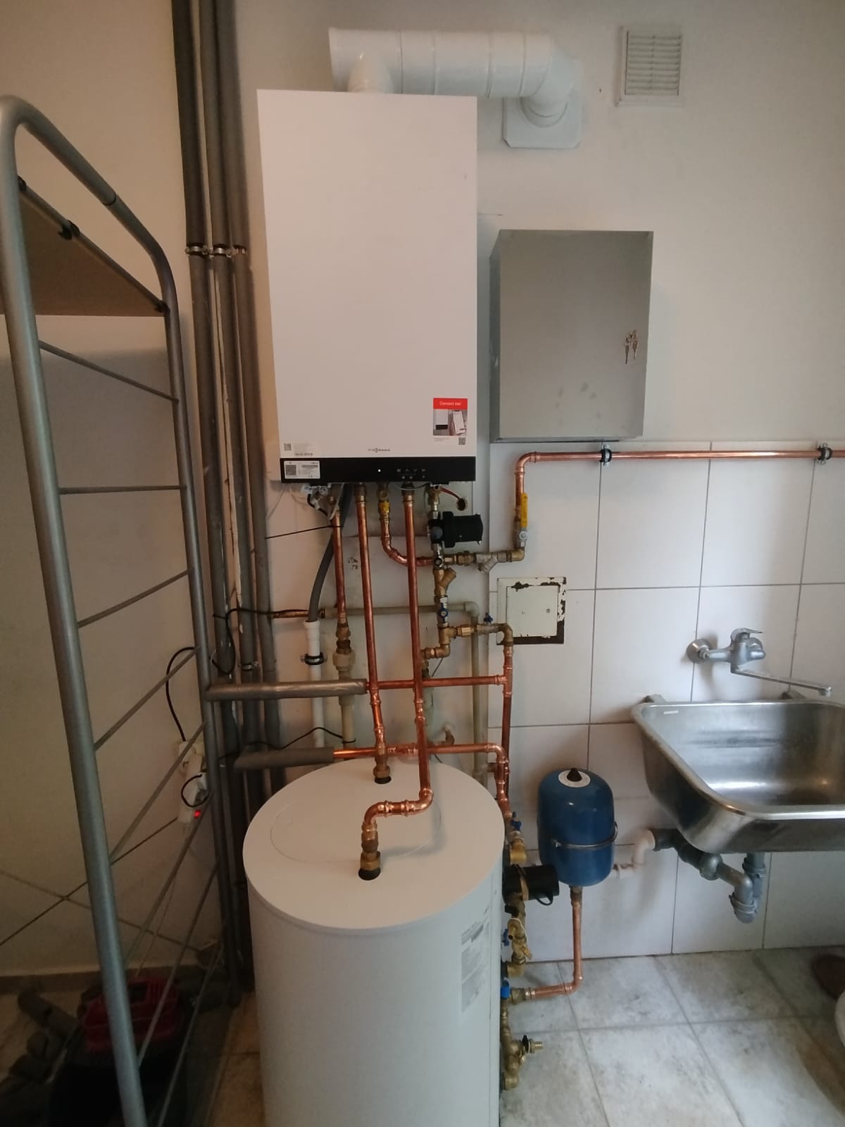 Biały piec gazowy Viessmann z miedzianymi rurami i zasobnikiem wody w pomieszczeniu z białymi płytkami. Widoczna instalacja i drabina.