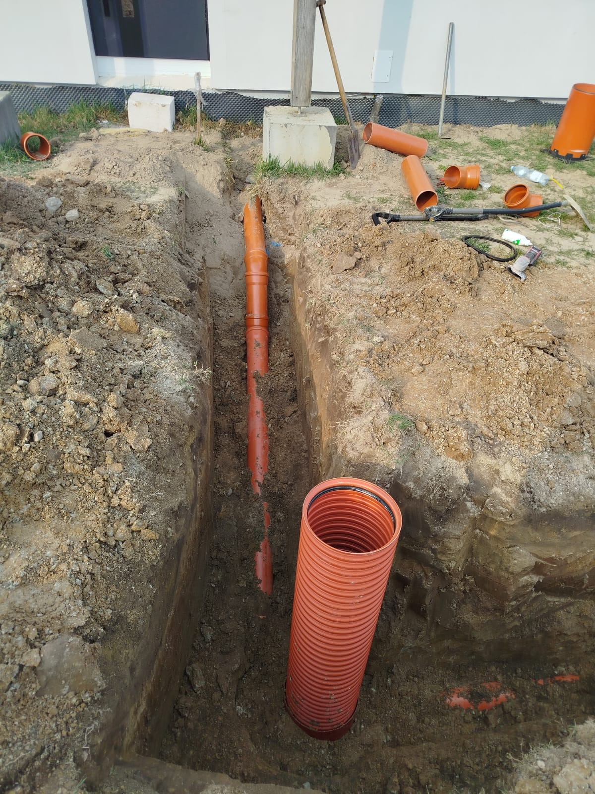 Wykonany wykop pod instalację rur kanalizacyjnych. Pomarańczowe rury częściowo ułożone w wykopie, inne leżą obok. Widoczny teren budowy i narzędzia.