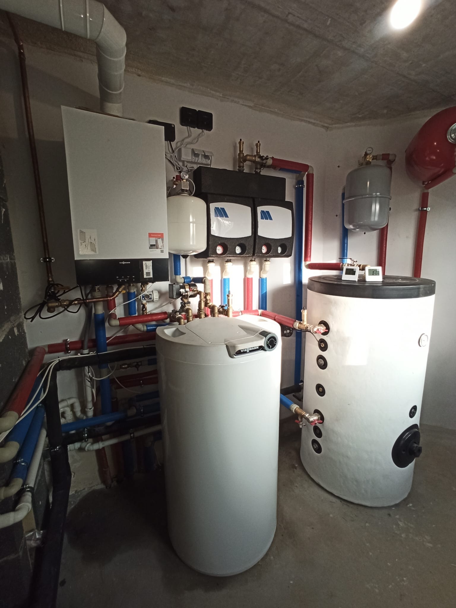 Kompletna kotłownia z dwoma zbiornikami, piecem i rozdzielaczami. Instalacja grzewcza z rurami w kolorach czerwonym i niebieskim, zamontowana w pomieszczeniu technicznym.