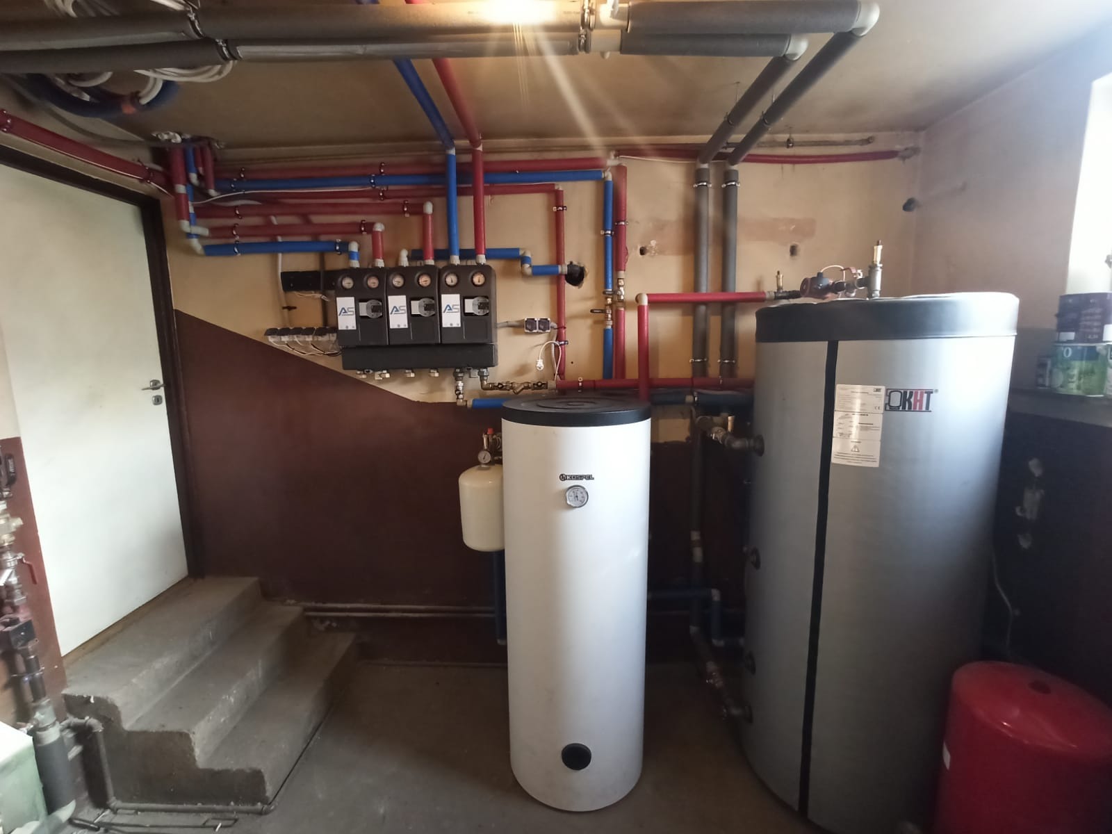 Kotłownia z dwoma zbiornikami, rurami w kolorach czerwonym i niebieskim oraz rozdzielaczami na ścianie. Widoczne schody i drzwi.