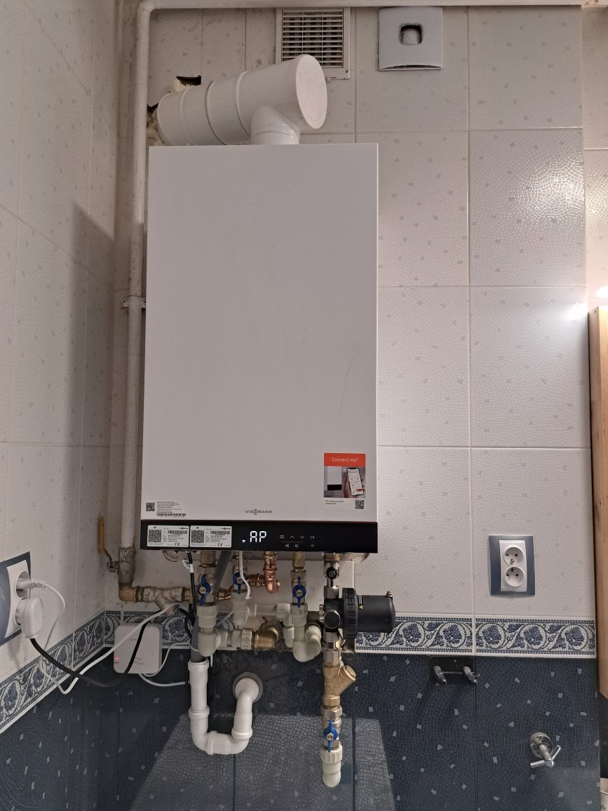 Nowoczesny piec gazowy Viessmann zamontowany na ścianie z dekoracyjnymi płytkami, widoczne liczne rury i zawory, gniazdko elektryczne po lewej stronie.