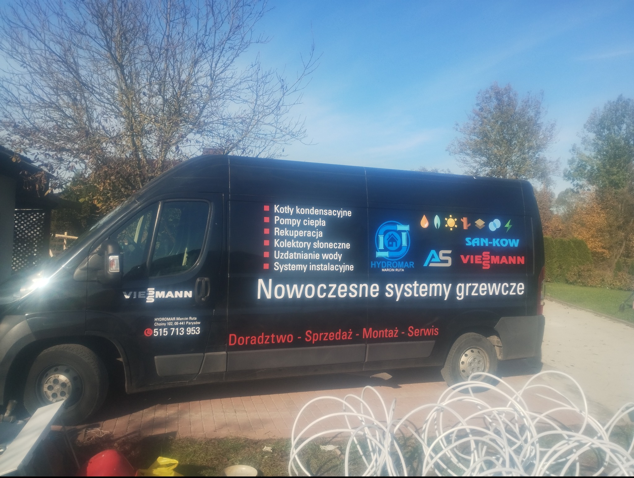 Czarny bus firmy Hydromar z reklamą nowoczesnych systemów grzewczych, w tle drzewa i trawnik. Na pierwszym planie rury do ogrzewania podłogowego.