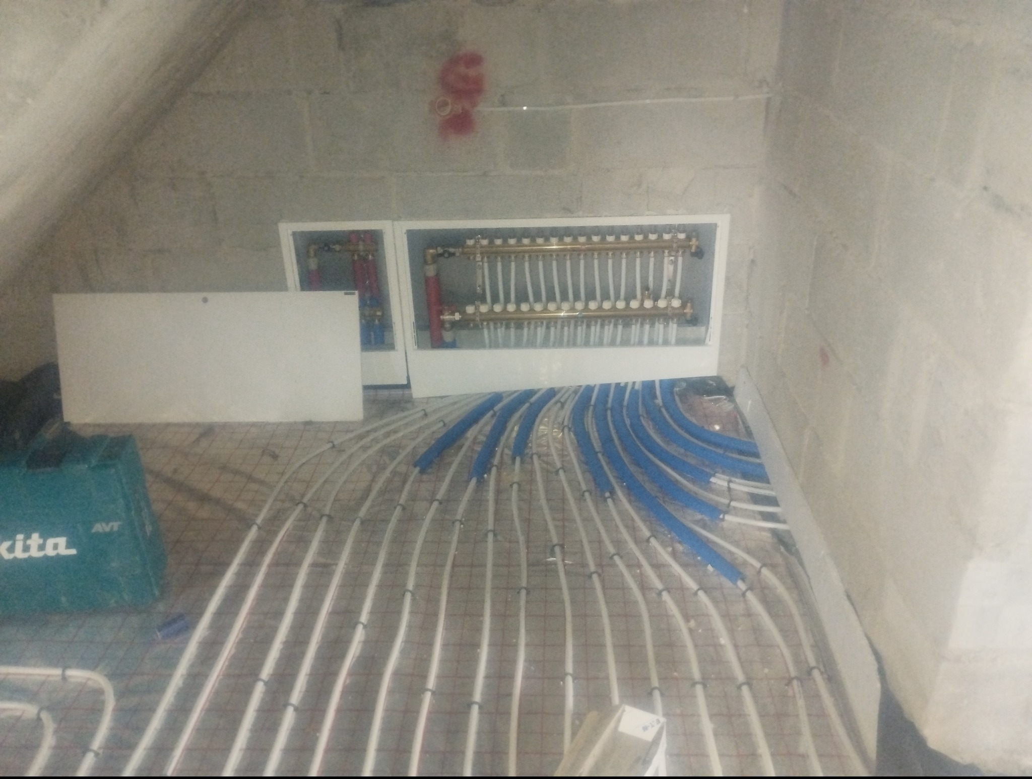 Rozłożone białe rury ogrzewania podłogowego na siatce, z niebieskimi elementami mocującymi. W tle rozdzielacz w białej obudowie na szarej ścianie.