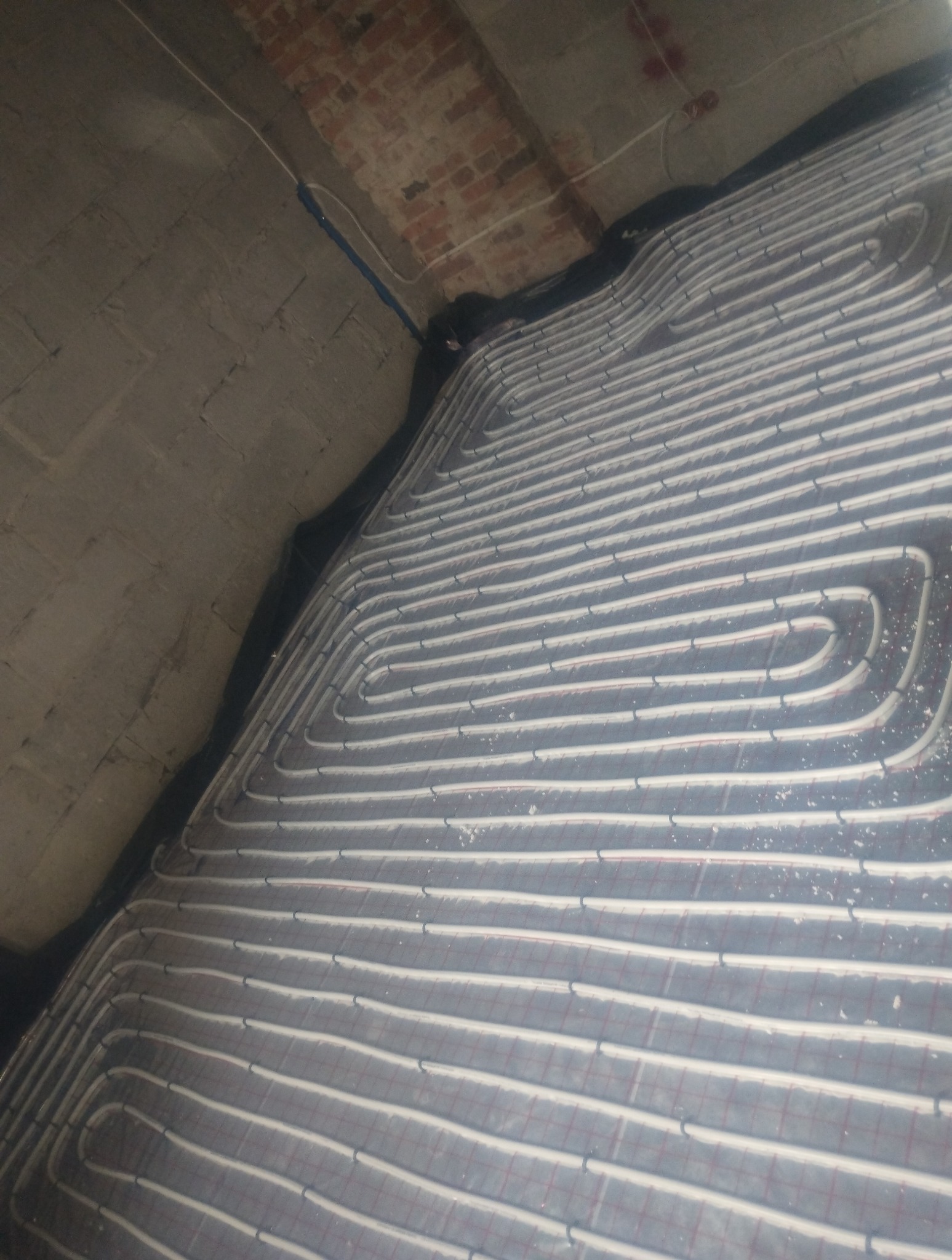 Szare rury ogrzewania podłogowego ułożone spiralnie na czarnej macie izolacyjnej, widoczne na tle surowego betonowego stropu i fragmentu ceglanego muru.