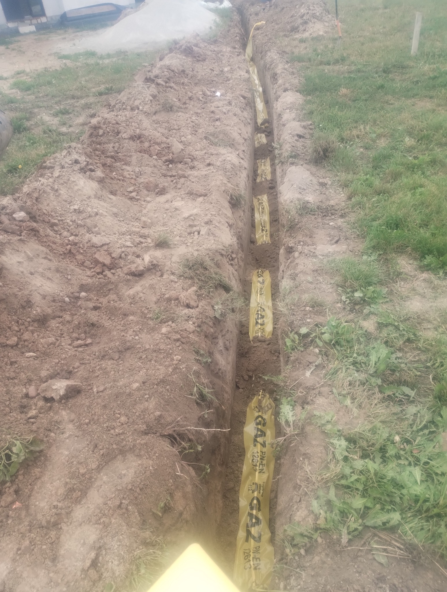 Wykop z żółtą folią oznaczającą gaz, widok z góry. Etap prac przyłącza gazowego na działce, ziemia, trawa.