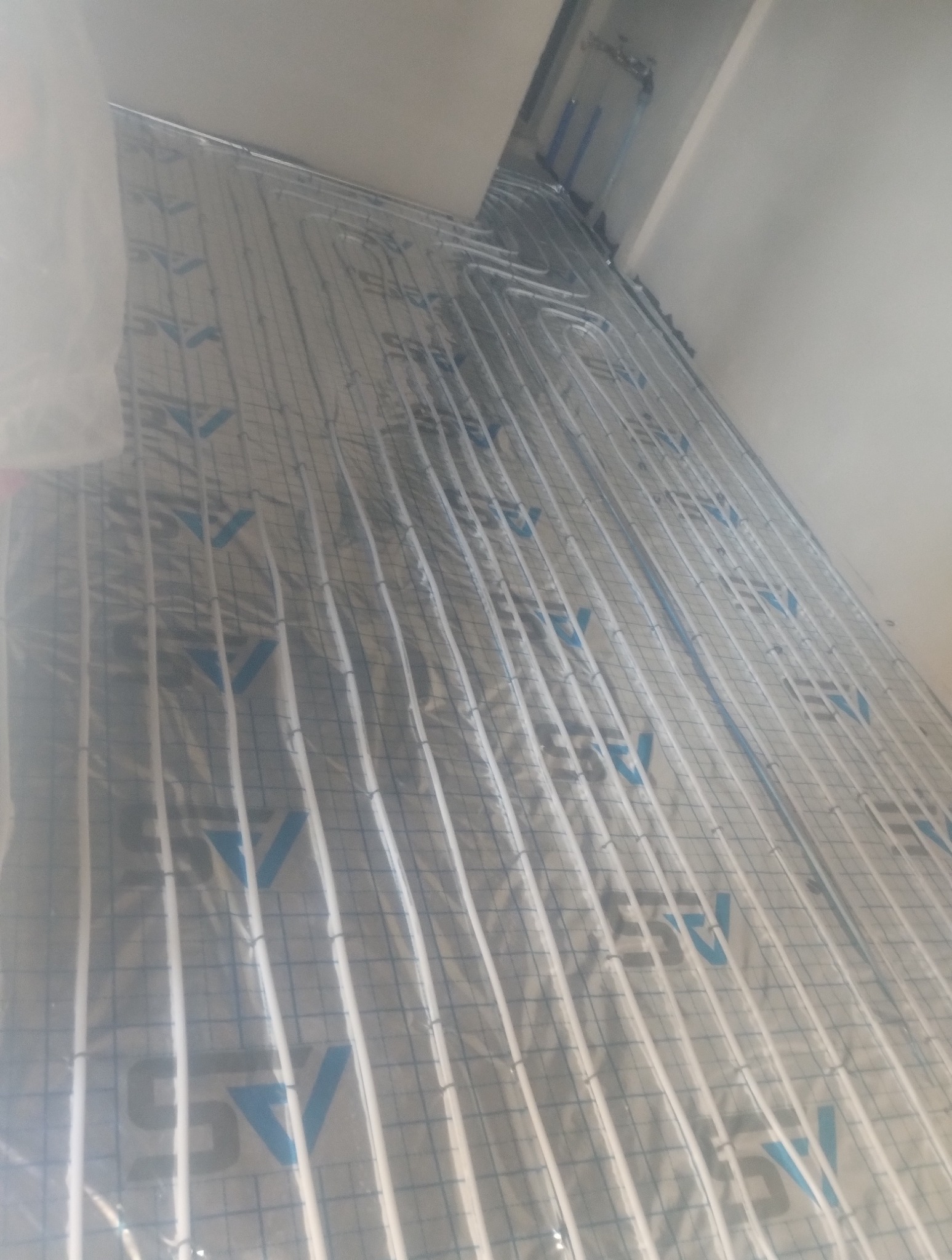 Białe rurki ogrzewania podłogowego ułożone na srebrnej folii z niebieskim nadrukiem, widok z góry, instalacja w trakcie realizacji.