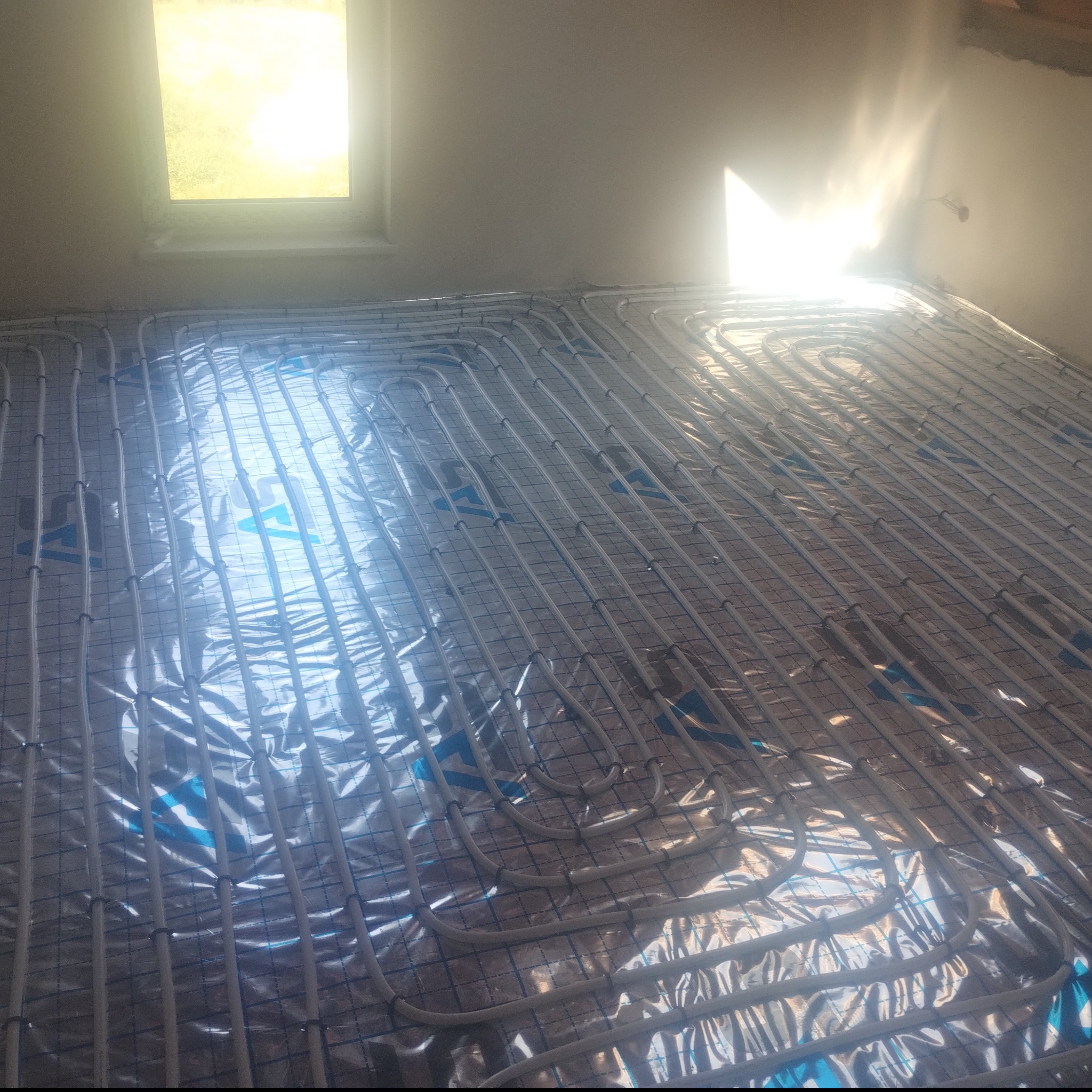 Ułożone rury ogrzewania podłogowego na folii izolacyjnej w jasnym pomieszczeniu z oknem. Widok z góry ukazuje regularny wzór instalacji.