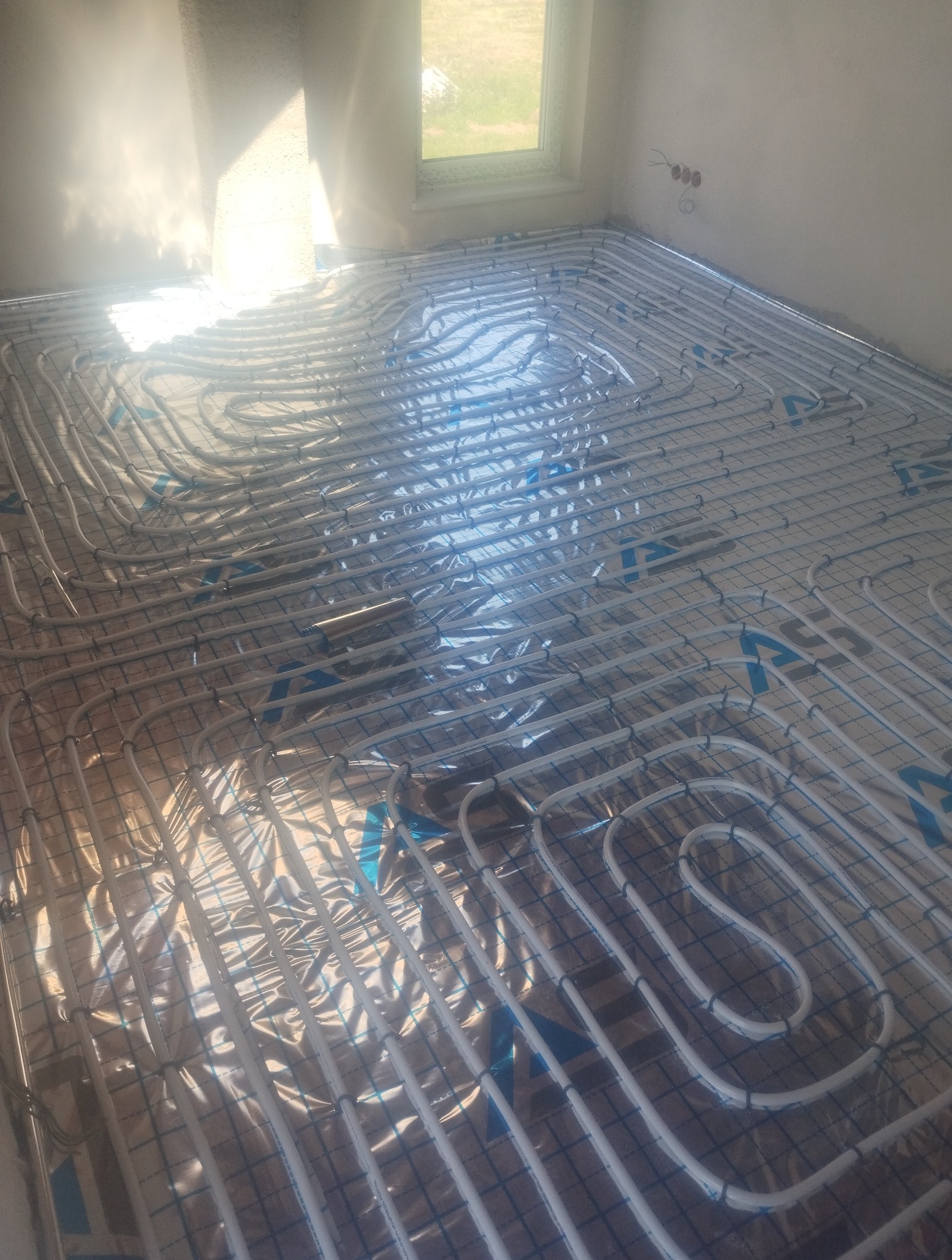 Białe rury ogrzewania podłogowego ułożone na folii izolacyjnej w jasnym pomieszczeniu z oknem. Widoczne mocowania rur. Realizacja przed wylaniem posadzki.