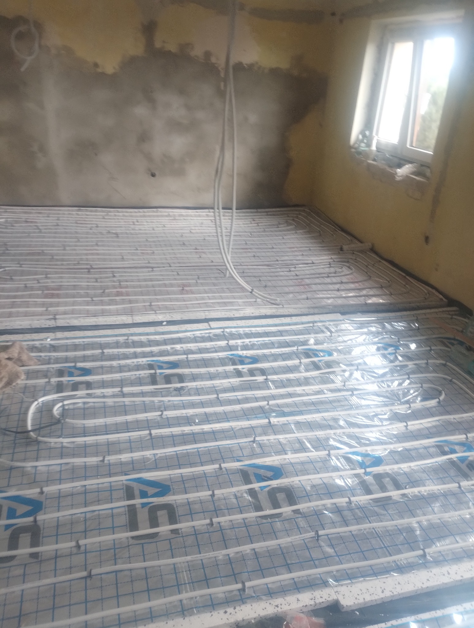 Białe rury ogrzewania podłogowego ułożone na izolacji w surowym pomieszczeniu z oknem, gotowe do zalania posadzką. Widoczne mocowania rur.