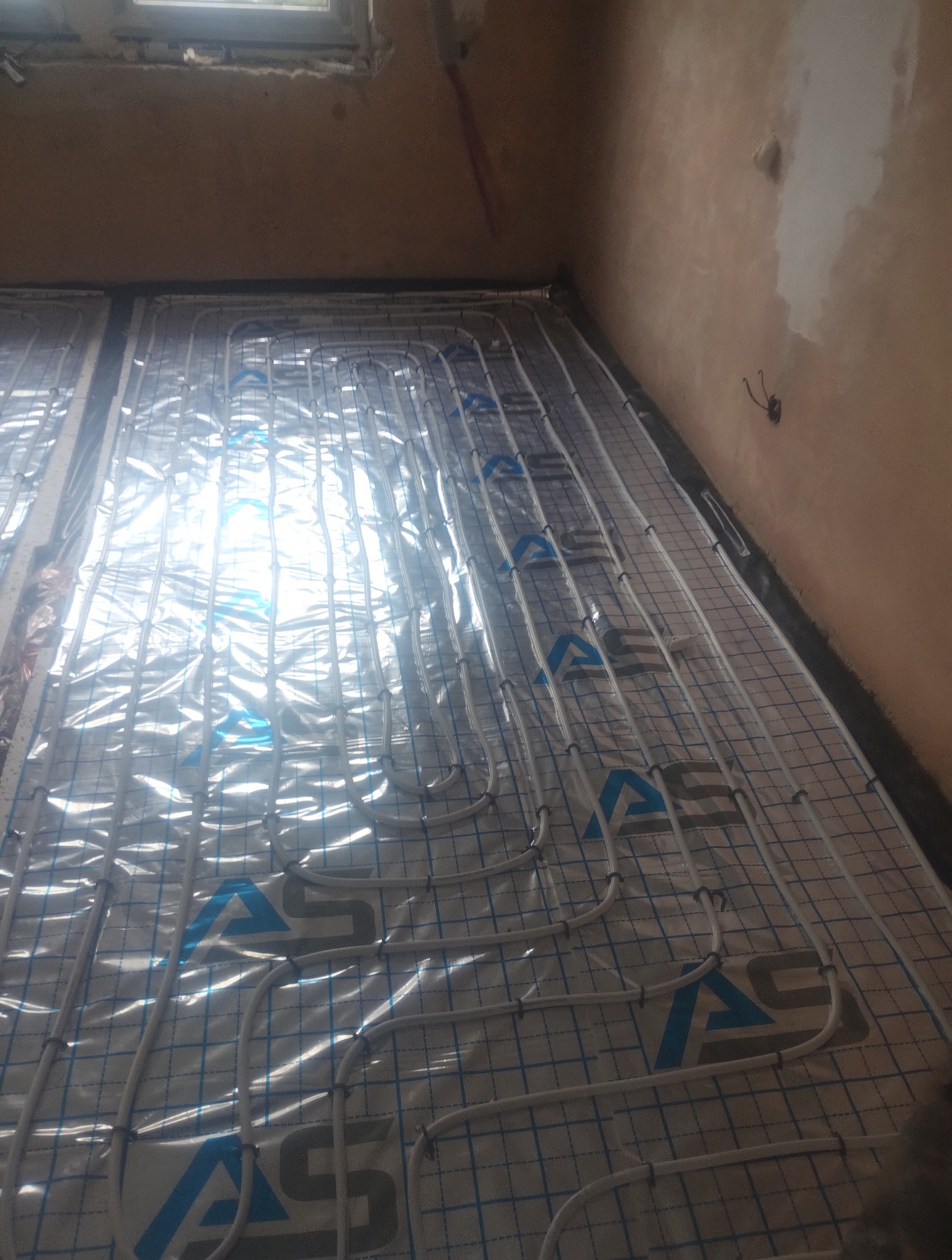 Białe rury ogrzewania podłogowego ułożone na folii z nadrukiem, zamocowane do siatki. Widok na fragment ściany i okno. Instalacja w trakcie realizacji.