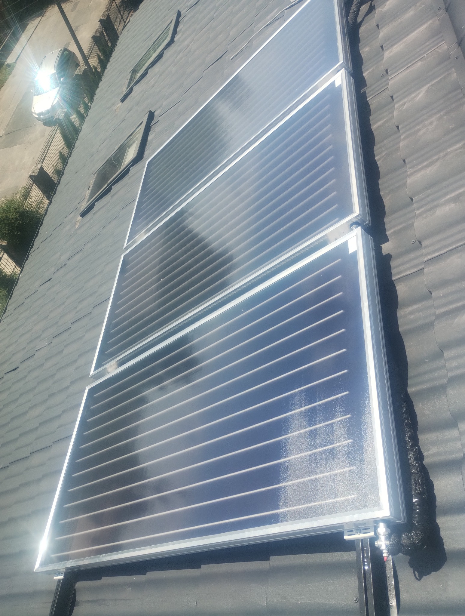 Ukończony montaż kolektorów słonecznych na dachu pokrytym szarą dachówką. Widoczny fragment instalacji i okna dachowe. Ujęcie z góry, ostre światło słoneczne.