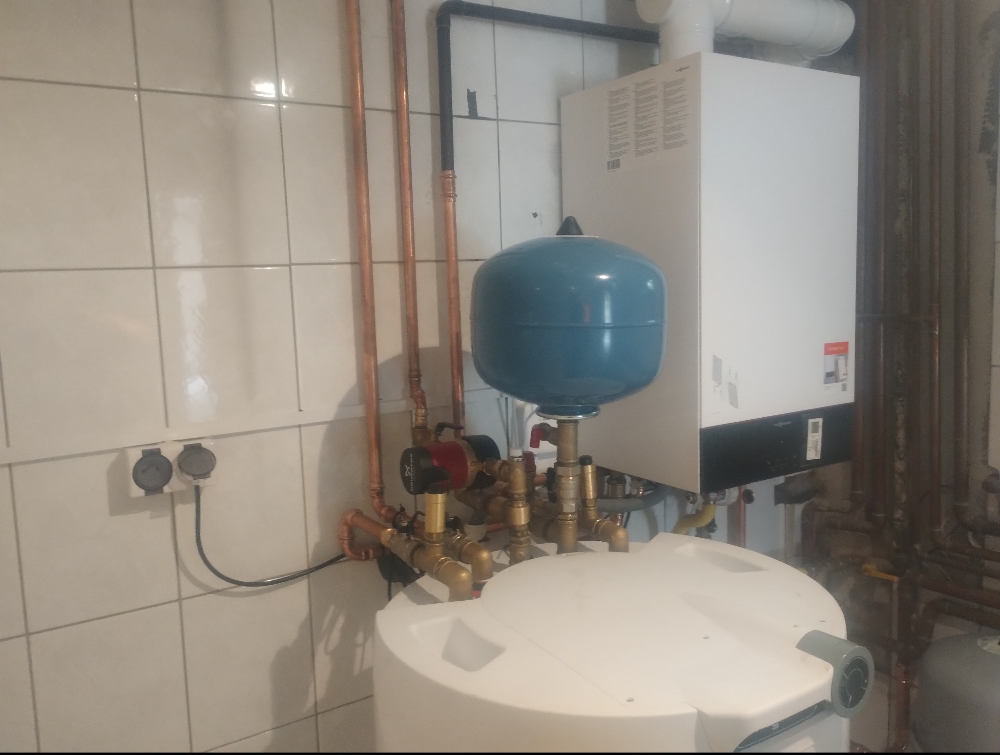 Zmontowana instalacja CO z piecem gazowym, zbiornikiem i rurami miedzianymi na tle białych, kwadratowych płytek. Widoczne przyłącza elektryczne i elementy armatury.