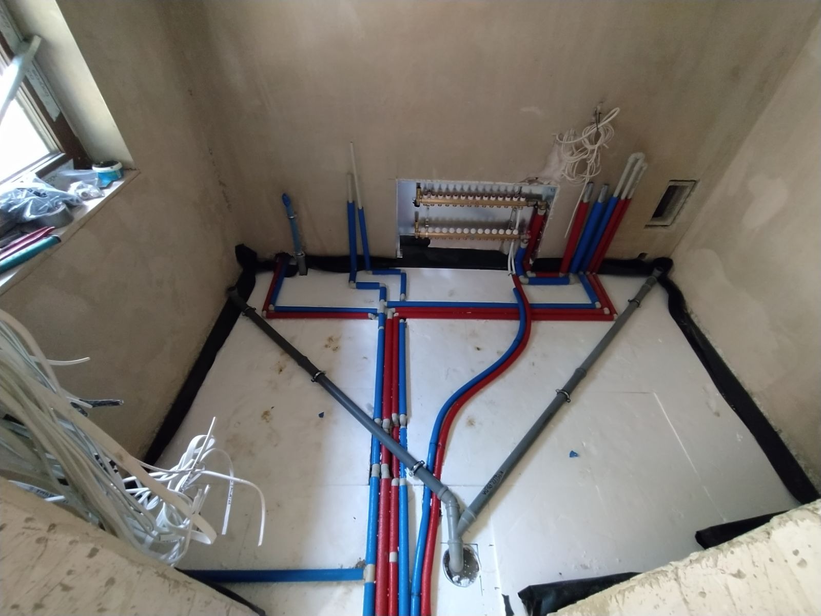 Rozłożone rury instalacji ogrzewania podłogowego w surowym pomieszczeniu, widoczne elementy rozdzielacza i izolacji. Ujęcie z góry.