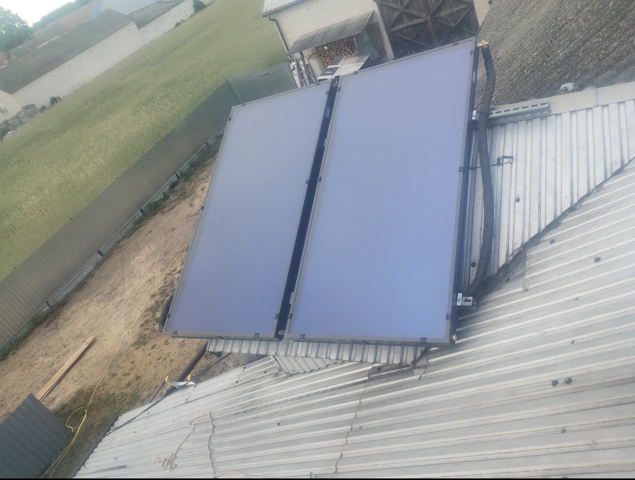 Dwa panele solarne zamontowane na blaszanym dachu budynku, widoczne przewody instalacji. W tle trawnik i budynki gospodarcze.