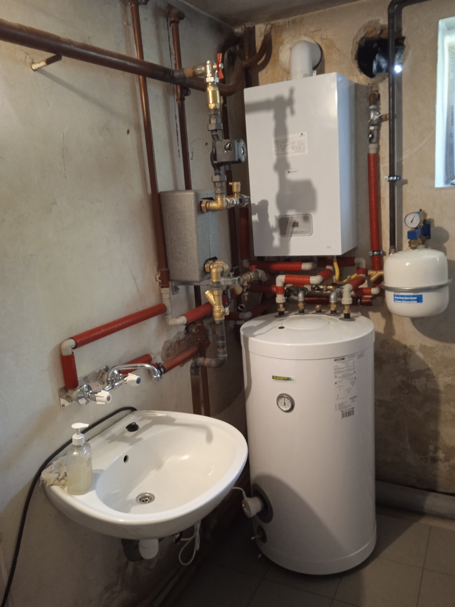 Biały piec CO z podłączonymi rurami miedzianymi i zbiornikiem wyrównawczym, zamontowany w piwnicy obok umywalki. Widoczne liczne zawory i elementy instalacji.