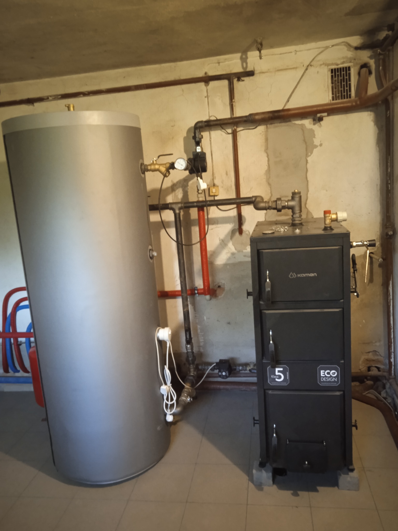 Szary bojler i czarny piec marki Kamen z podłączeniami w kotłowni. Widoczne rury i elementy instalacji grzewczej.