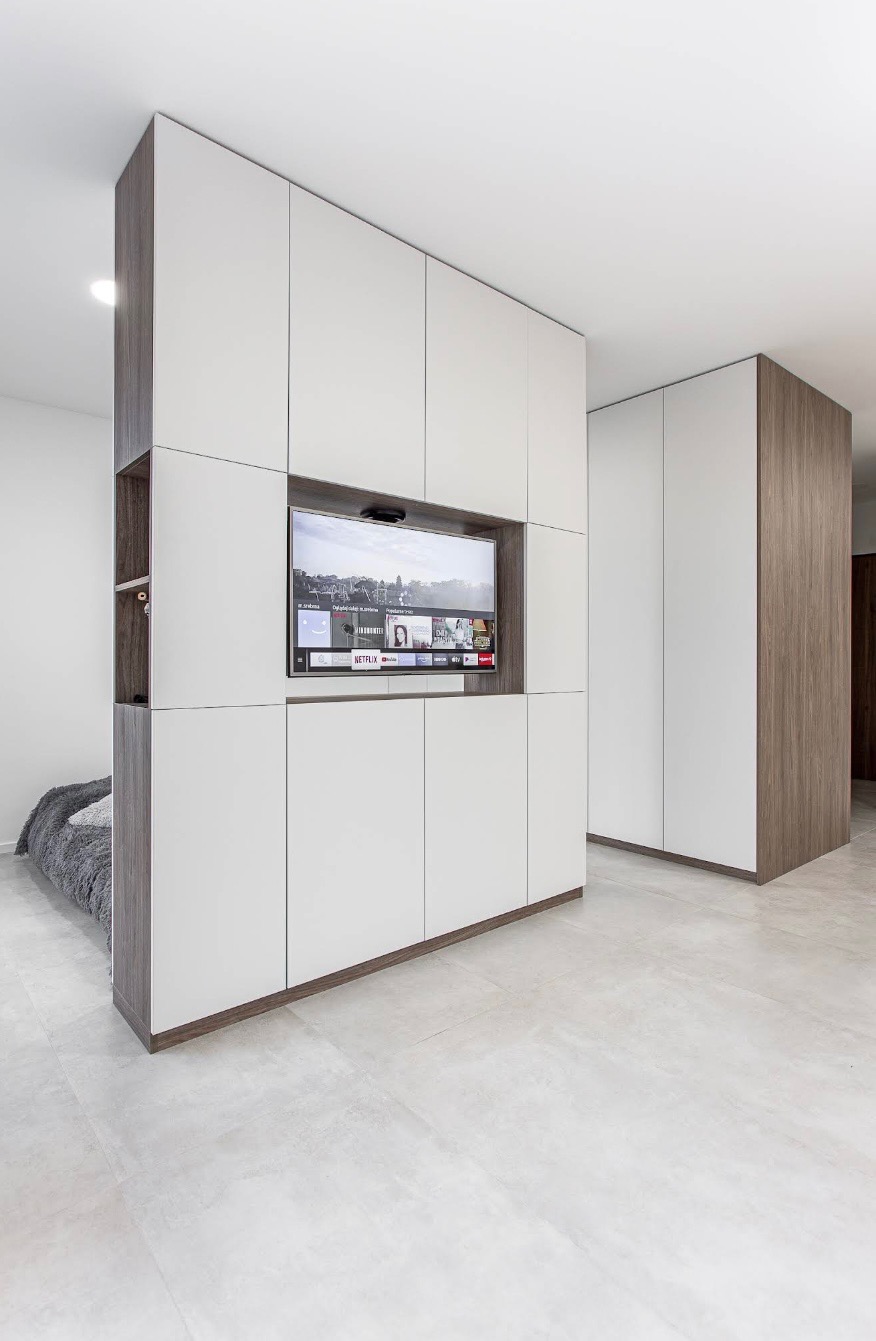 Nowoczesna zabudowa meblowa z białymi frontami i drewnianymi akcentami, integrująca telewizor. Minimalistyczny design z szafkami i półkami w pokoju.