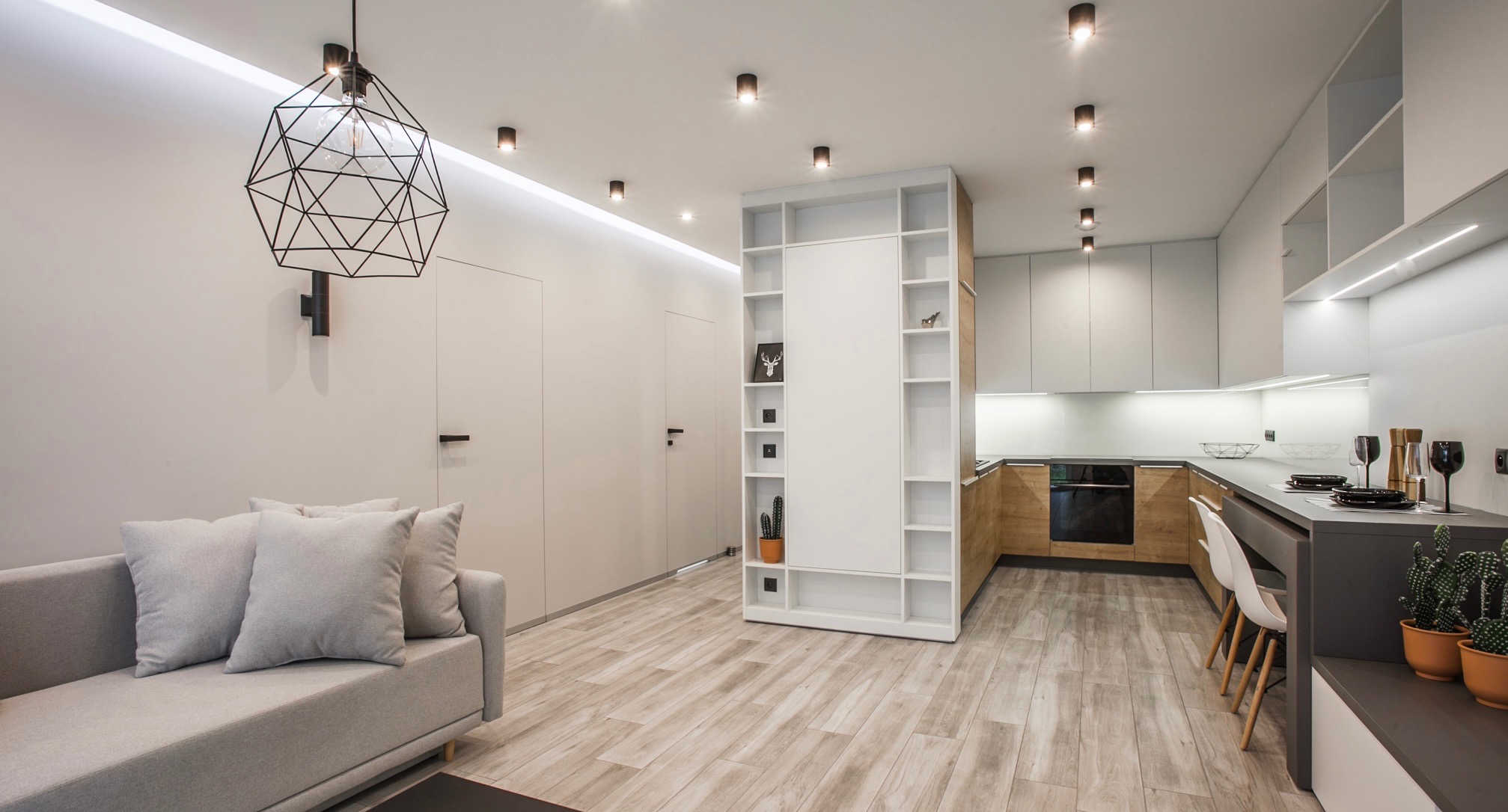 Nowoczesne wnętrze: szara sofa z poduszkami, geometryczna lampa, białe drzwi i regał, drewniana podłoga, kuchnia z drewnianymi i szarymi szafkami, oświetlenie punktowe.