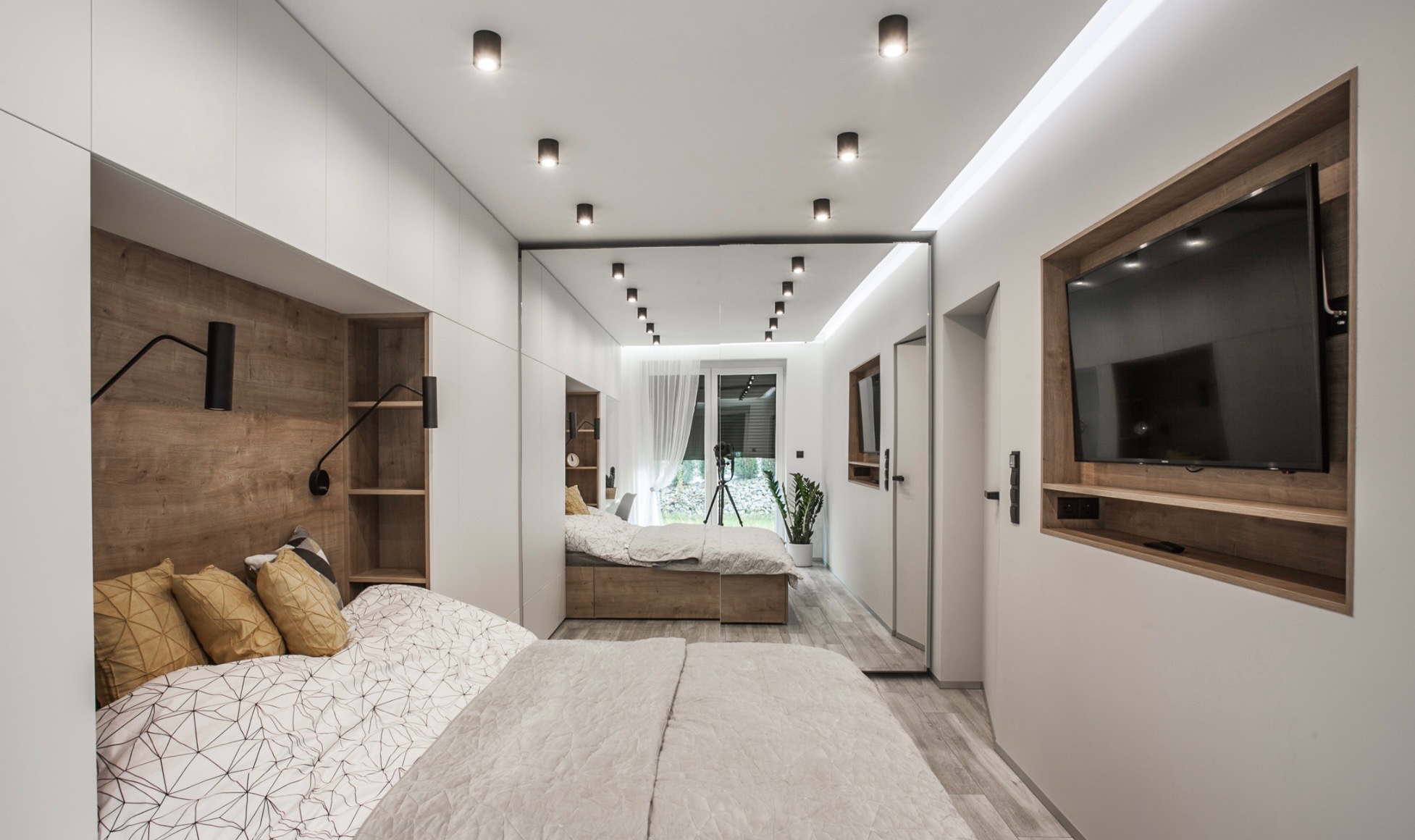 Nowoczesna sypialnia z lustrzaną ścianą, wbudowaną szafą i telewizorem. Drewniane akcenty, designerskie lampki nocne i minimalistyczny wystrój tworzą przytulną atmosferę.