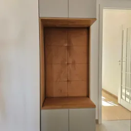 Funkcjonalna zabudowa wnęki z tapicerowanym siedziskiem i szafkami w odcieniach szarości i brązu, widok na jasne wnętrze przez otwarte drzwi.