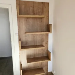 Drewniane półki ścienne z płyty wiórowej w odcieniach brązu, zamocowane na ścianie w pokoju, widok z boku, minimalistyczny design, widoczne gniazdko elektryczne.