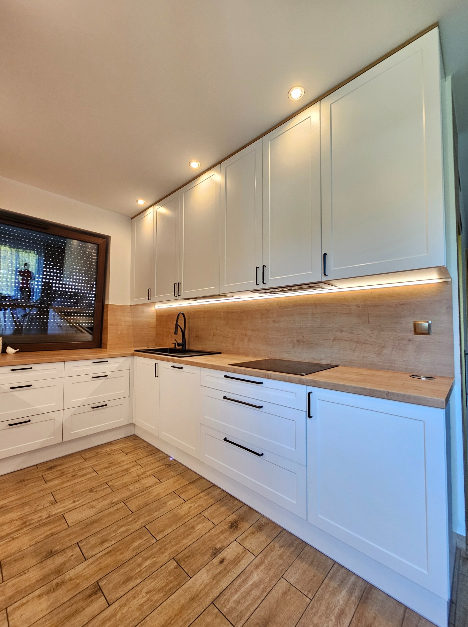 Biała kuchnia z drewnianymi blatami i czarną armaturą, oświetlona listwami LED pod szafkami wiszącymi i punktowymi lampami w suficie. Widoczna płyta indukcyjna.