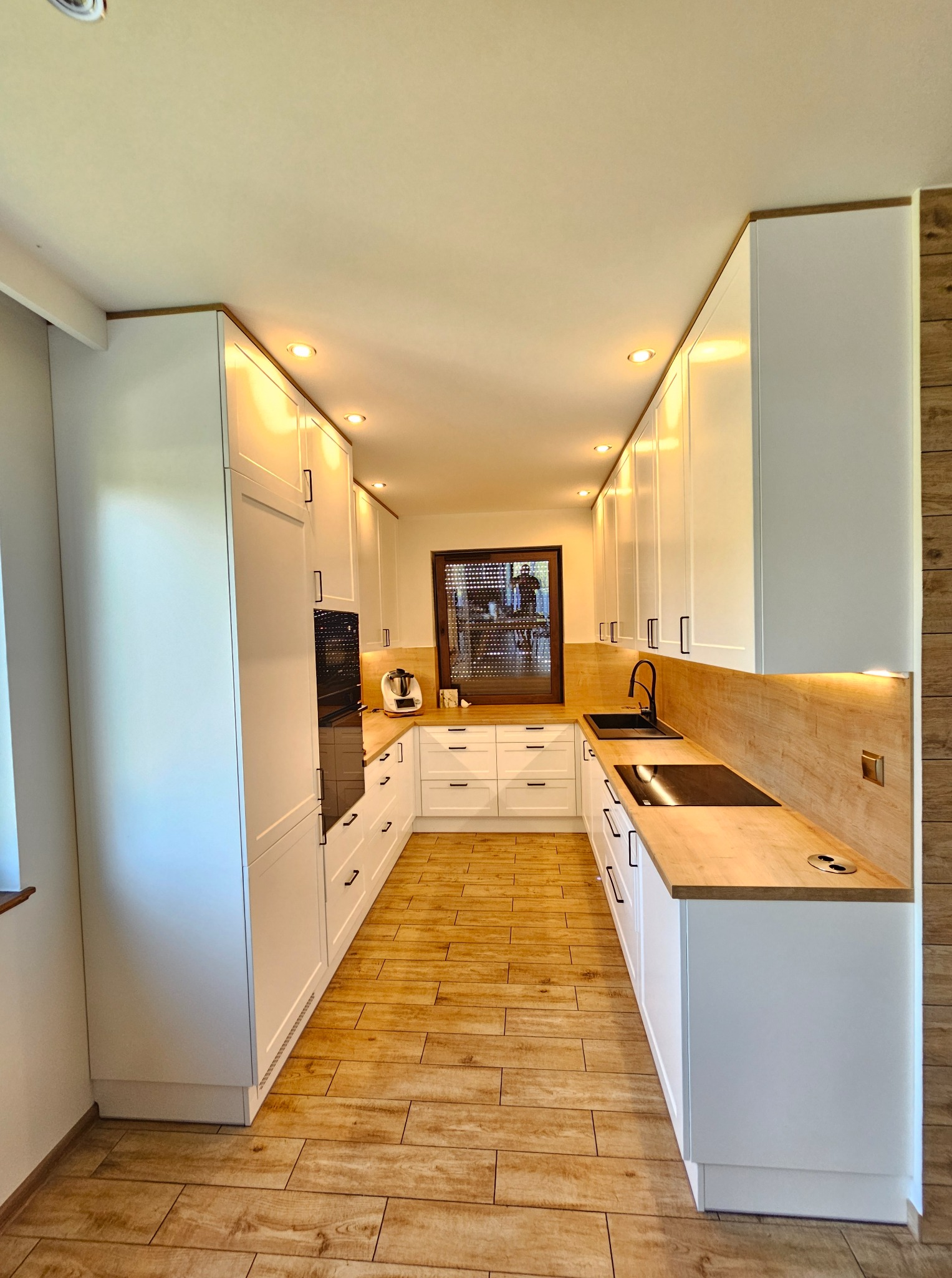 Nowoczesna kuchnia w kształcie litery U z białymi frontami, drewnianym blatem i podświetleniem LED. Widok na okno z roletami i sprzęt AGD.