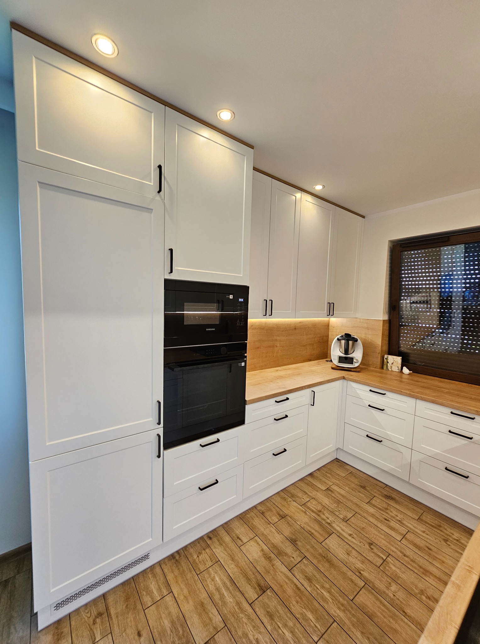 Jasna kuchnia z białymi szafkami i drewnianym blatem. Uwagę przykuwają podłużne, czarne uchwyty oraz zabudowany piekarnik i płyta grzewcza.