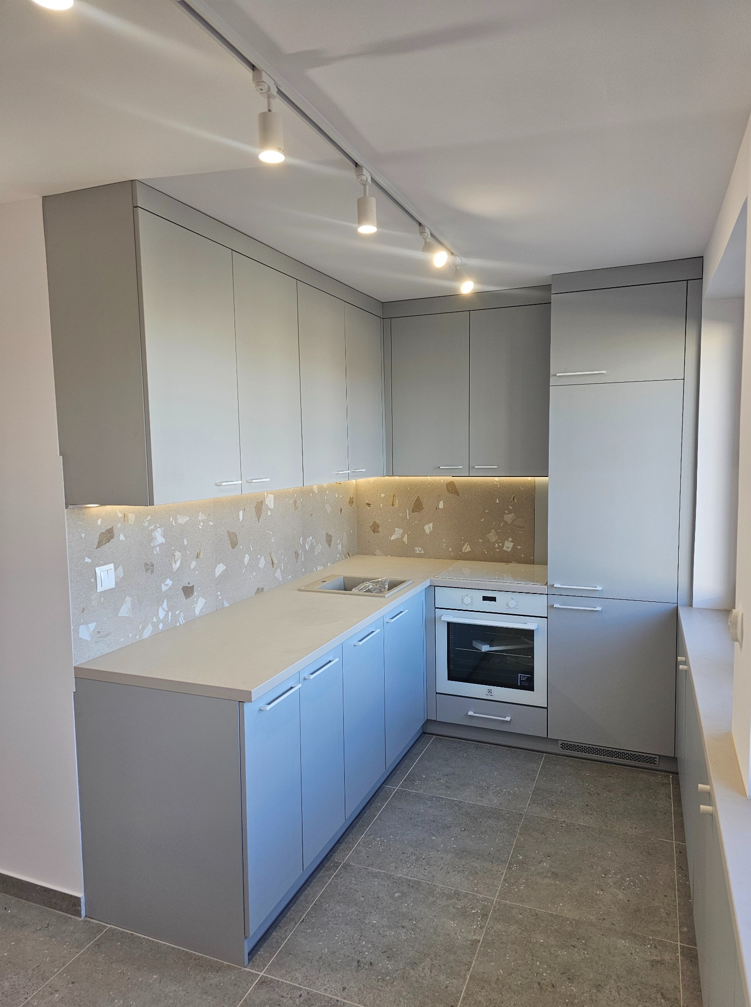 Jasnoszara kuchnia z blatem w odcieniach szarości i beżu, oświetlona listwą LED pod szafkami wiszącymi i reflektorami sufitowymi. Uwagę zwraca wzór na ścianie.