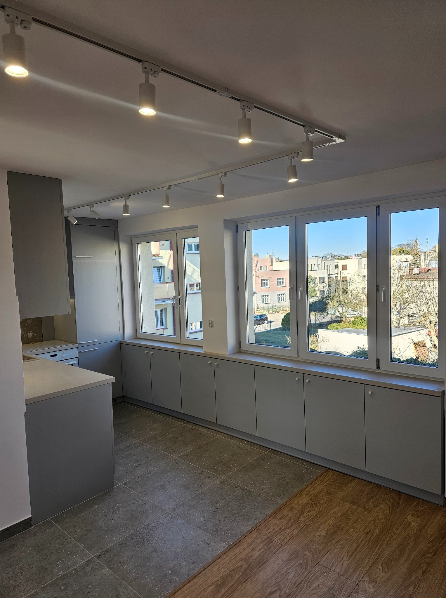 Szara kuchnia z długim blatem pod oknem i oświetleniem szynowym. Minimalistyczny design, widok na budynki za oknem. Nowoczesne wykończenie.