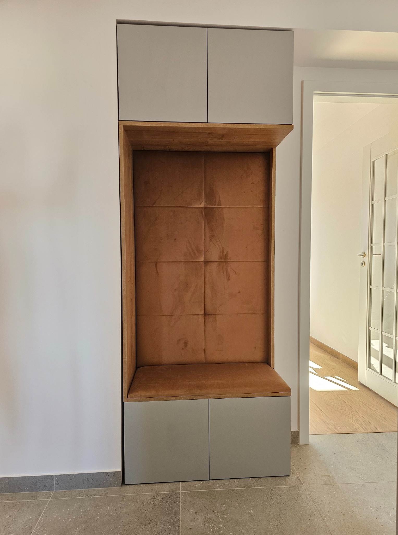 Funkcjonalna zabudowa wnęki z tapicerowanym siedziskiem i szafkami w odcieniach szarości i brązu, widok na jasne wnętrze przez otwarte drzwi.