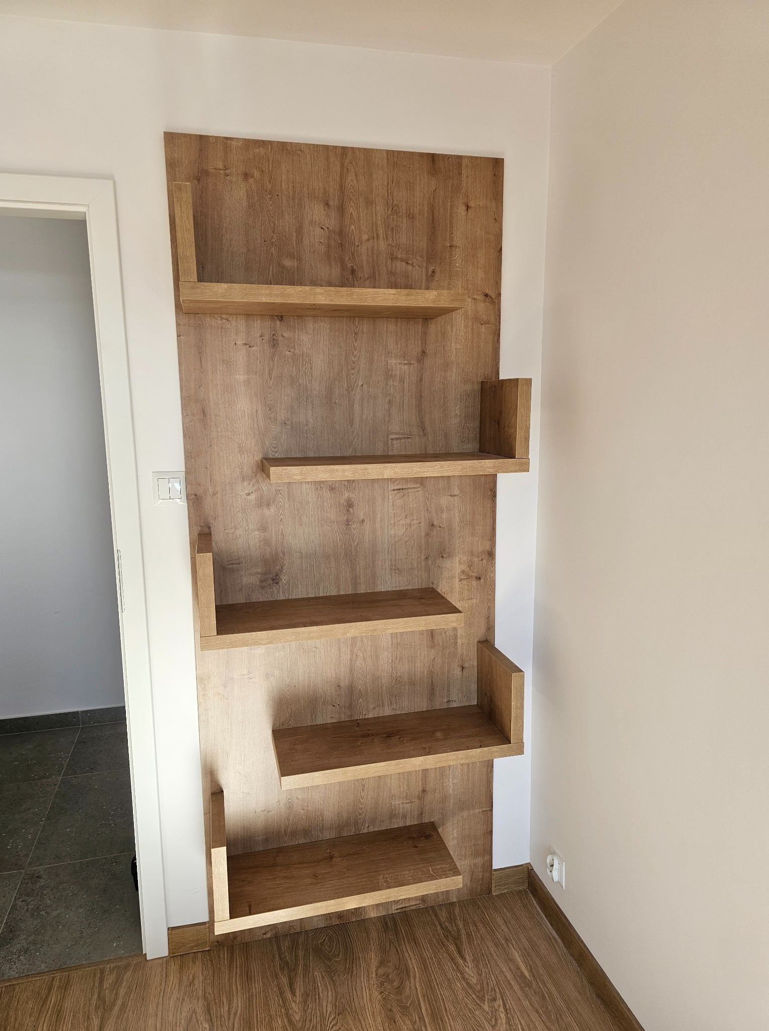 Drewniane półki ścienne z płyty wiórowej w odcieniach brązu, zamocowane na ścianie w pokoju, widok z boku, minimalistyczny design, widoczne gniazdko elektryczne.