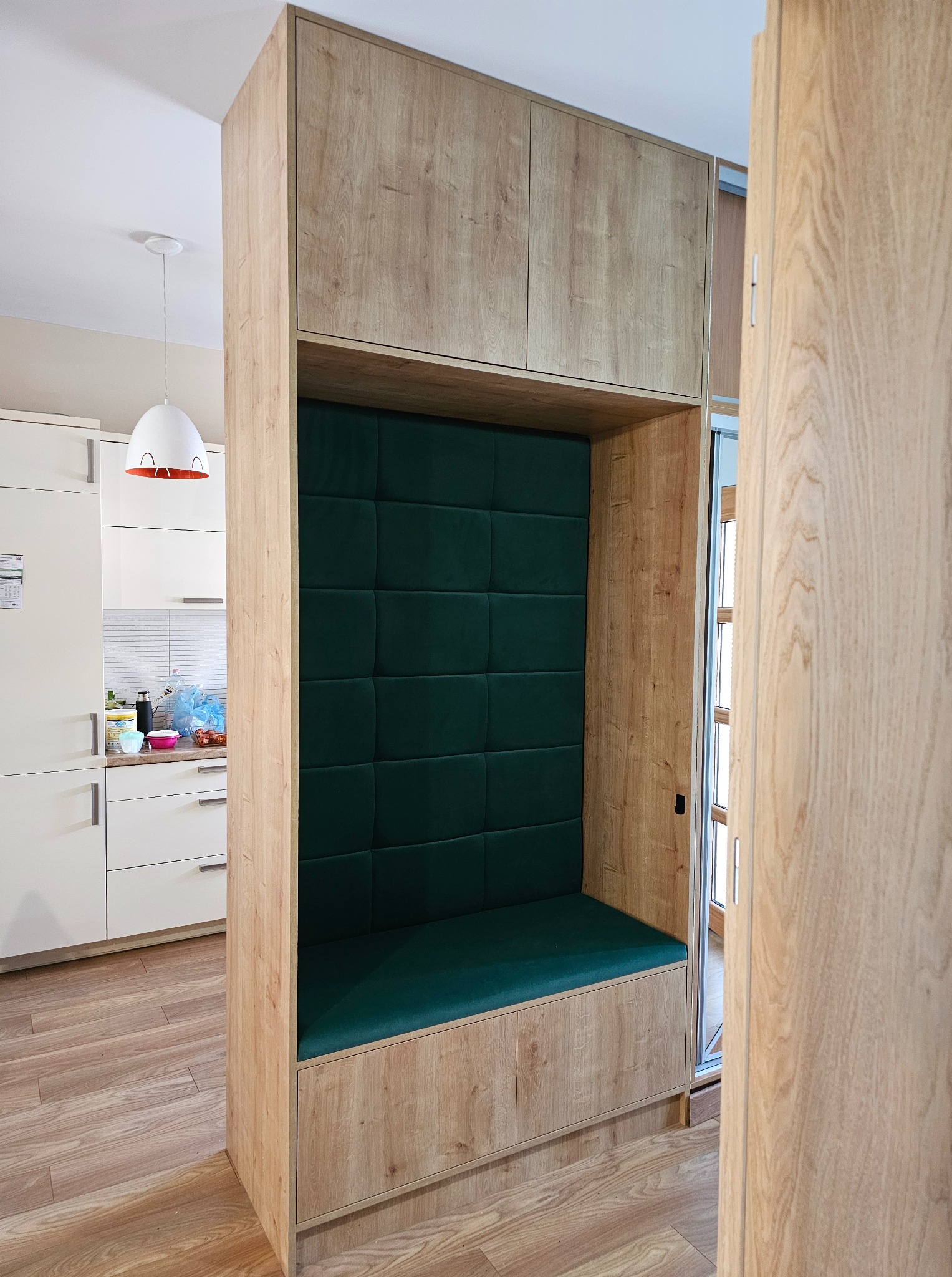 Wnęka z siedziskiem i zielonym obiciem, wbudowana w drewnianą zabudowę kuchenną z szafkami. Widok na białą kuchnię i podłogę z paneli.