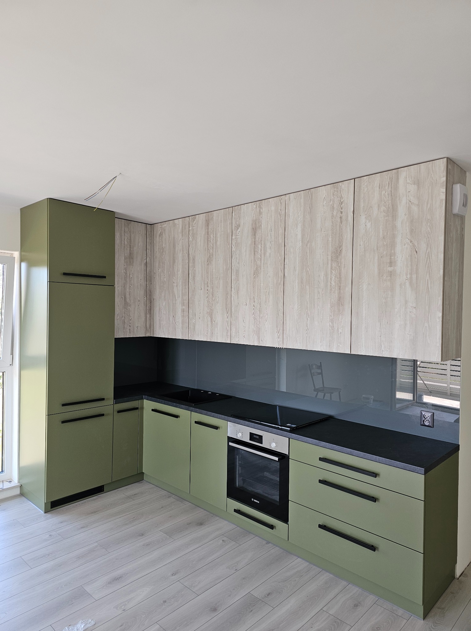 Nowoczesna kuchnia w odcieniach zieleni i drewna z czarnym blatem, srebrnym AGD i szklanym panelem. Widoczne gniazdka elektryczne.