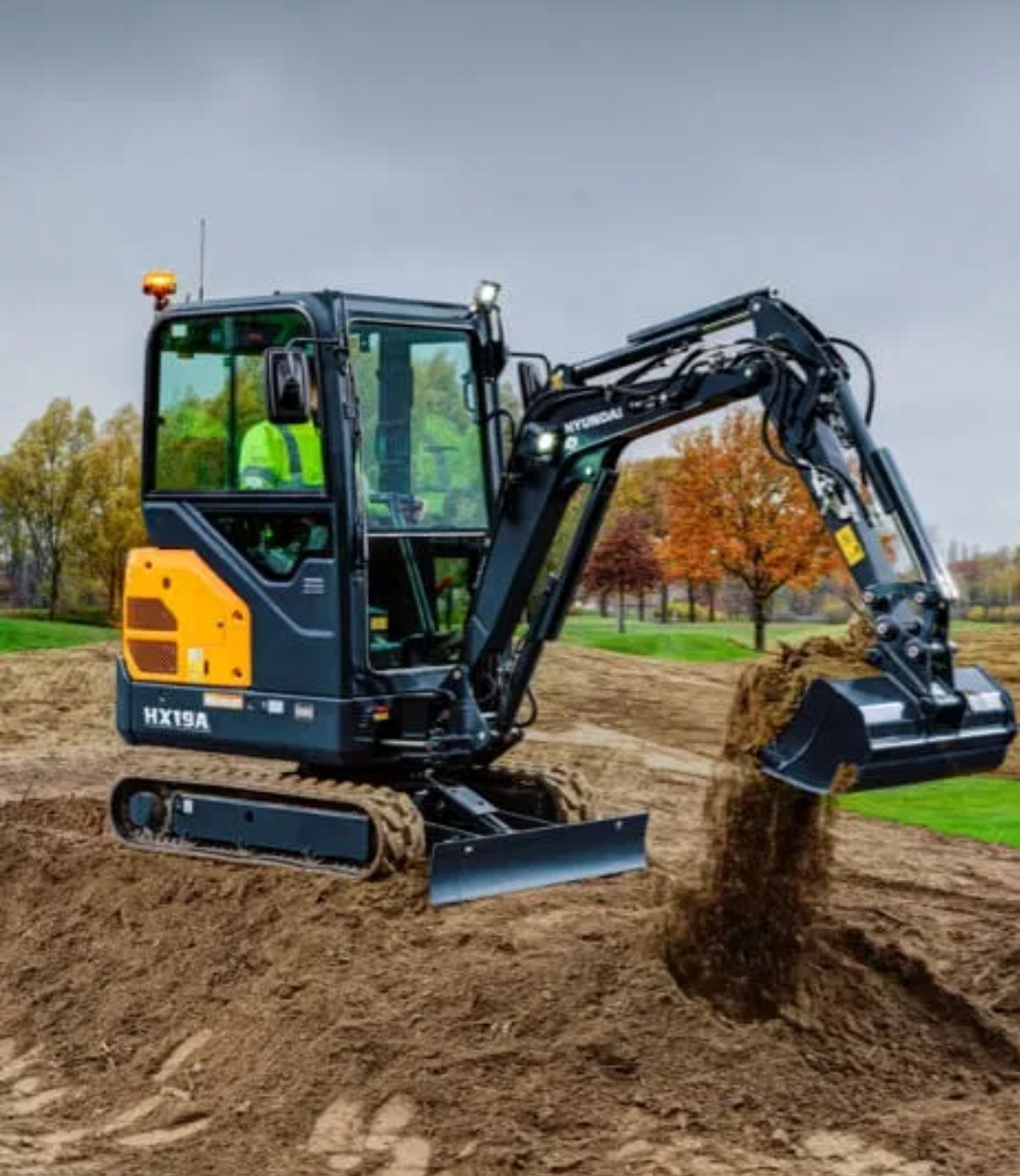 Mała koparka Hyundai HX19A w trakcie pracy, wysypująca ziemię z łyżki na polu. Operator w kamizelce odblaskowej widoczny w kabinie. Tło: drzewa z jesiennymi liśćmi.