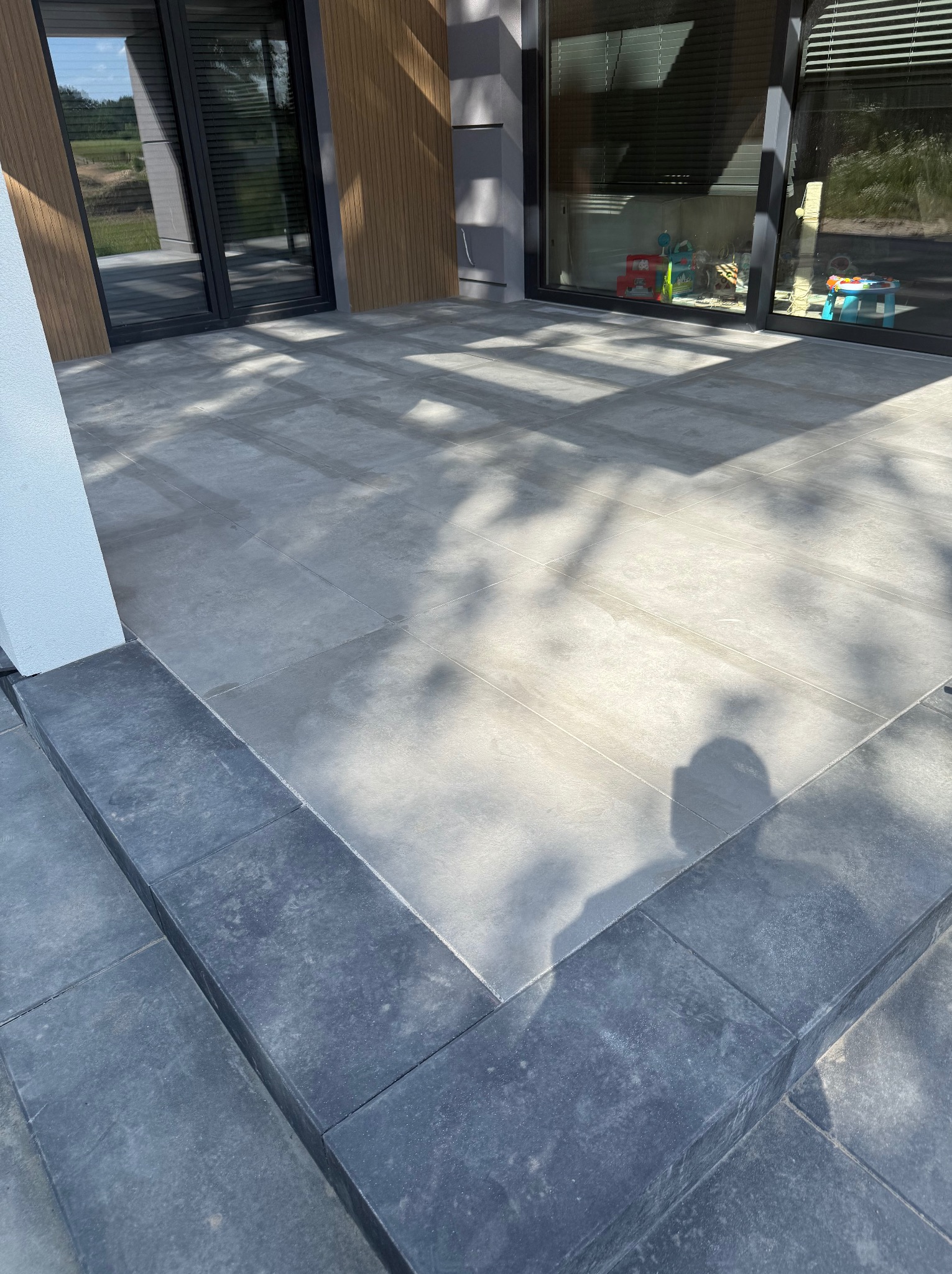 Nowoczesny taras z szarych, wielkoformatowych płyt. Widoczne detale łączeń i cień drzewa. Minimalistyczny design z elementami drewnianej elewacji w tle.