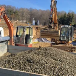 Żwirowy ładunek w skrzyni wywrotki, w tle dwie koparki Terex na placu budowy i wykop pod fundament. Błękitne niebo.