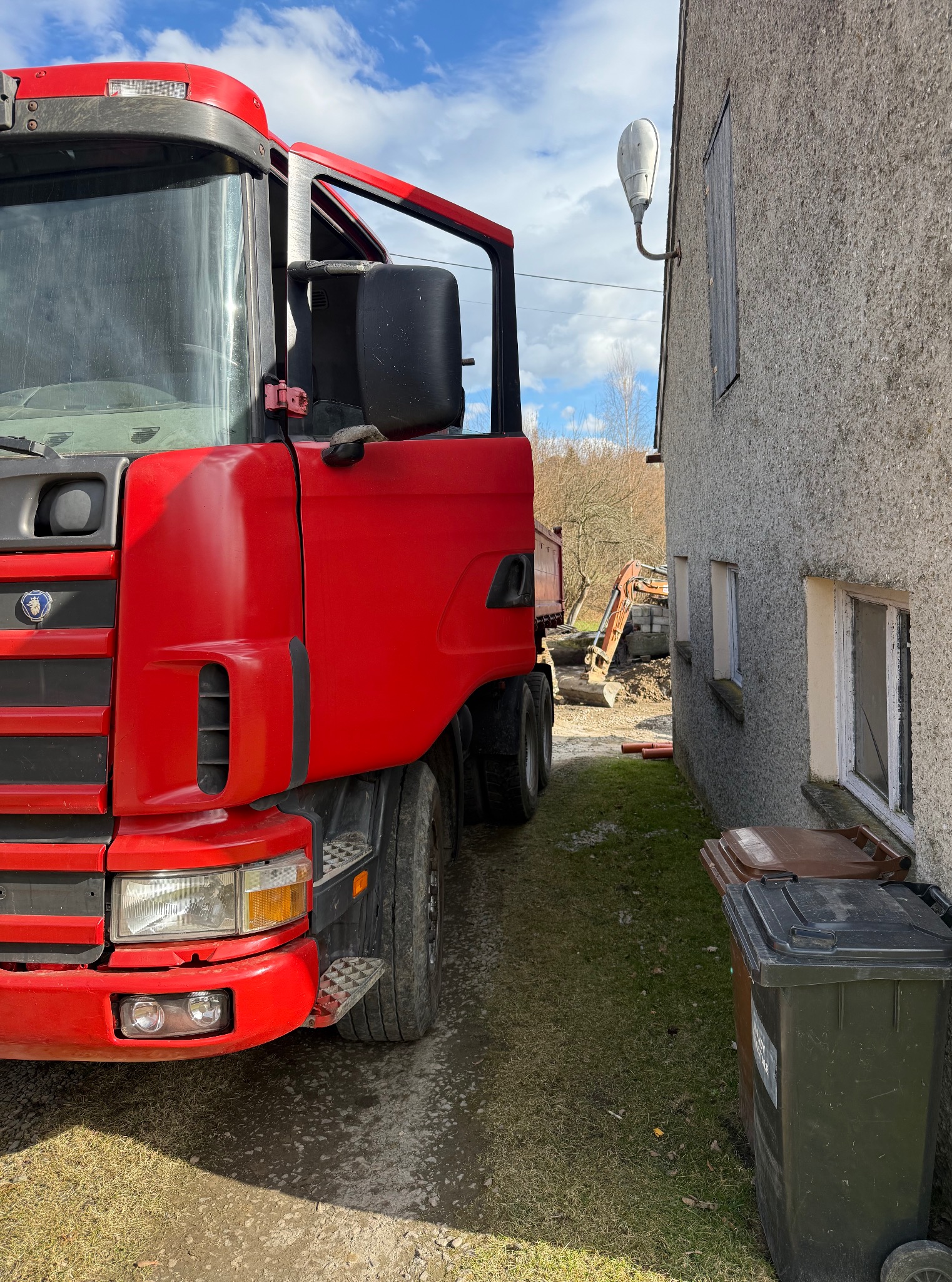 Czerwona ciężarówka Scania stoi obok budynku, w tle widoczna koparka przy wykopie. Błękitne niebo z chmurami. Kosze na śmieci przy budynku.