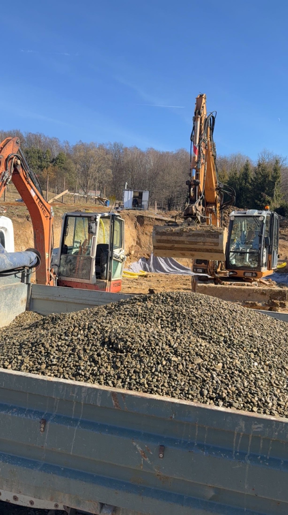 Żwirowy ładunek w skrzyni wywrotki, w tle dwie koparki Terex na placu budowy i wykop pod fundament. Błękitne niebo.
