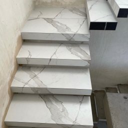 DV interiors - Wykończone schody z białych płytek imitujących marmur, z ciemnymi bokami, w surowym wnętrzu. Widok z góry.