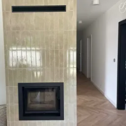 Obudowa kominka z beżowych, błyszczących płytek, czarny wkład kominkowy i kratki wentylacyjne. Parkiet w jodełkę, białe ściany, czarne futryny.