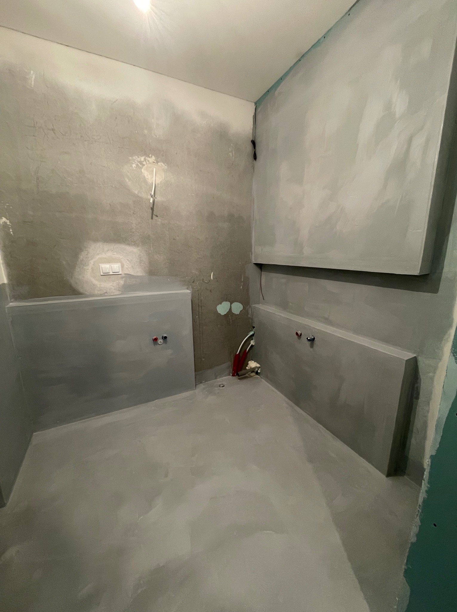 Surowa łazienka w szarościach: ściany z gładzią, przygotowane pod zabudowę, widoczne instalacje wodne. Minimalistyczny, nowoczesny styl w trakcie wykończenia.