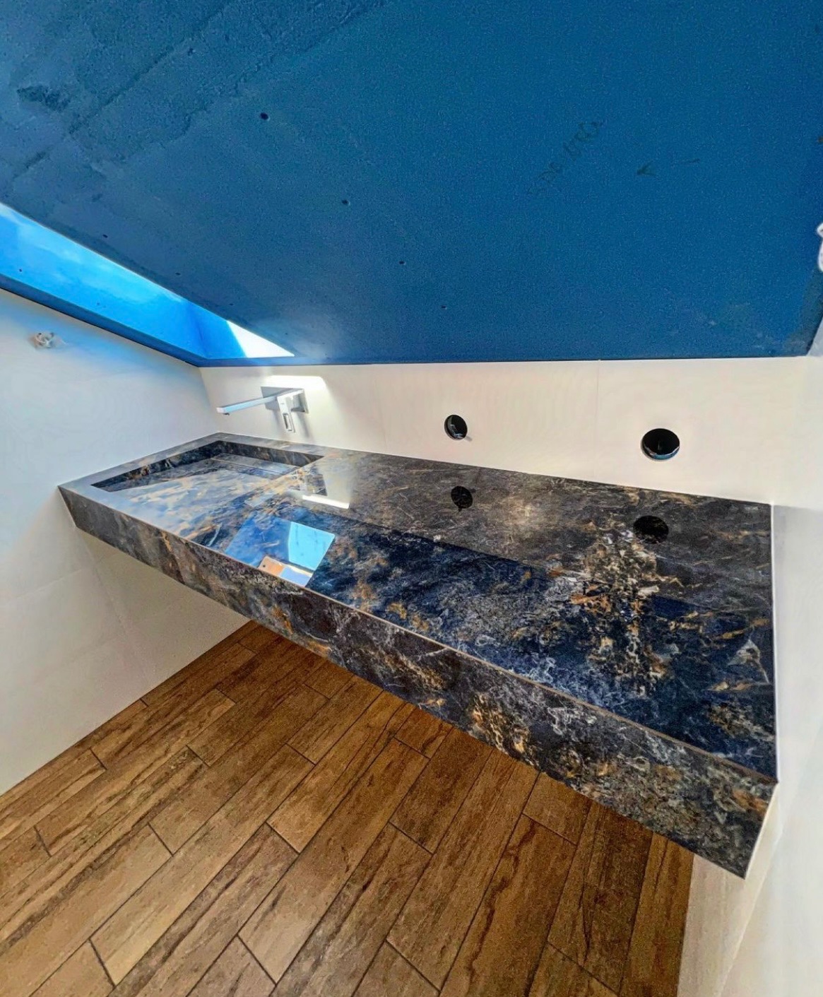 Wykończona łazienka: blat z ciemnego marmuru z umywalką pod oknem dachowym, drewnopodobna podłoga. Niebieski sufit i białe ściany tworzą nowoczesny kontrast.