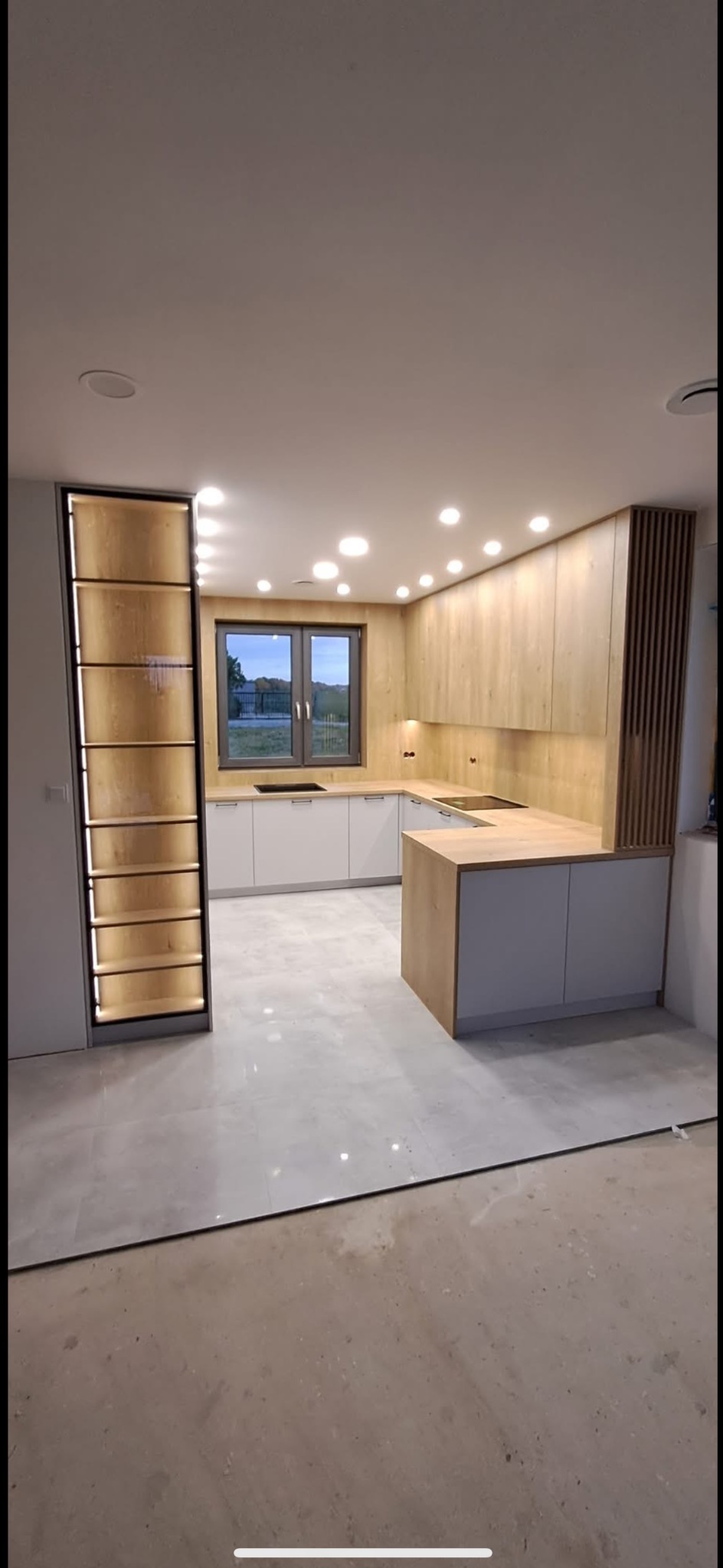 Nowoczesna kuchnia w odcieniach bieli i drewna z oświetleniem punktowym, widok na okno i częściowo wykończoną podłogą, prezentująca minimalistyczny design.