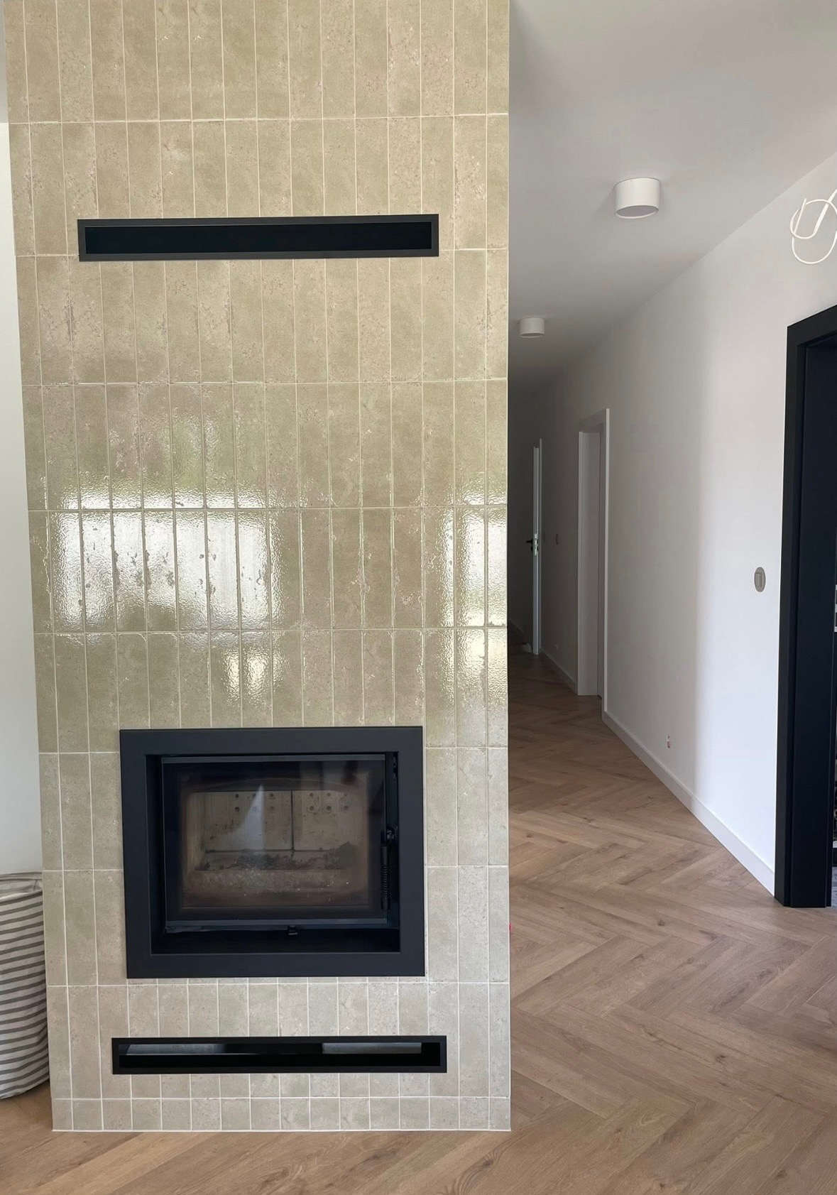 Obudowa kominka z beżowych, błyszczących płytek, czarny wkład kominkowy i kratki wentylacyjne. Parkiet w jodełkę, białe ściany, czarne futryny.