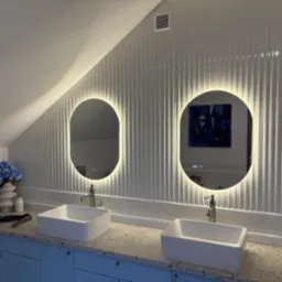 Nowoczesna łazienka na poddaszu z dwoma owalnymi lustrami LED, białymi umywalkami i niebieskimi szafkami. Ściany wyłożone białymi panelami.