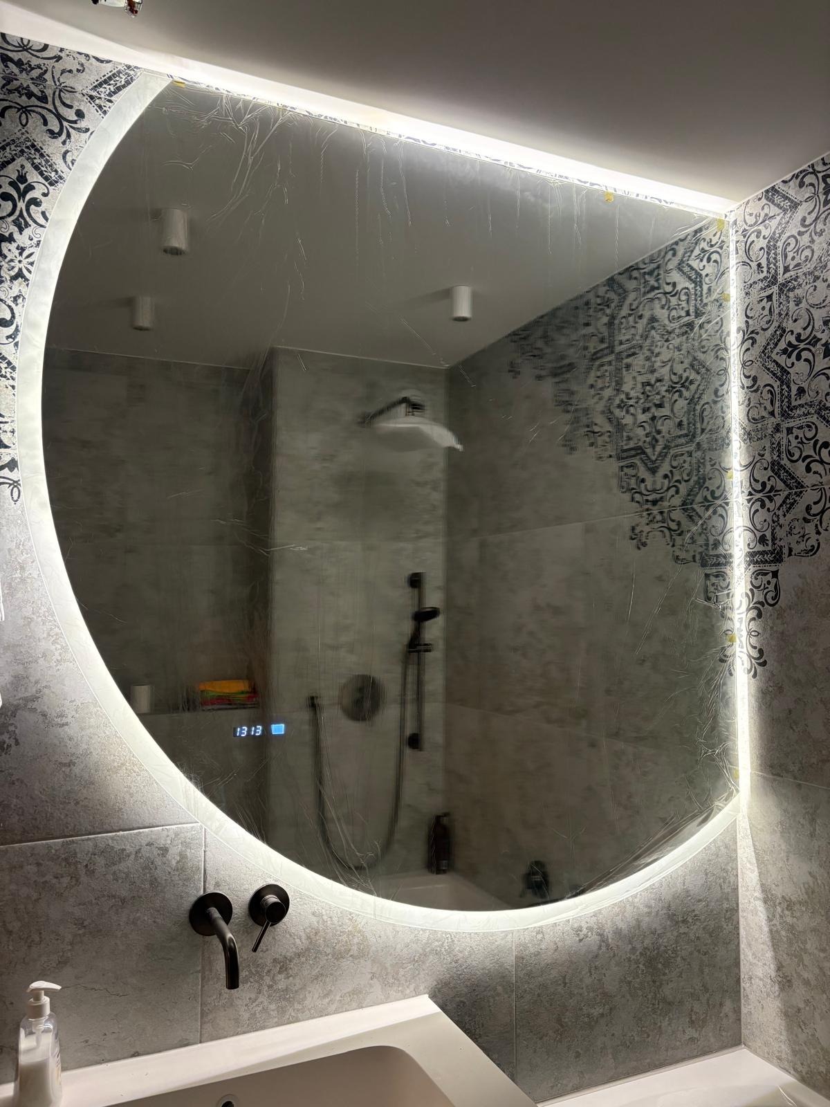 Okrągłe lustro LED w łazience z szarą mozaiką, widoczne elementy armatury i odbicie prysznica. Na lustrze folia ochronna, podświetlenie LED włączone.